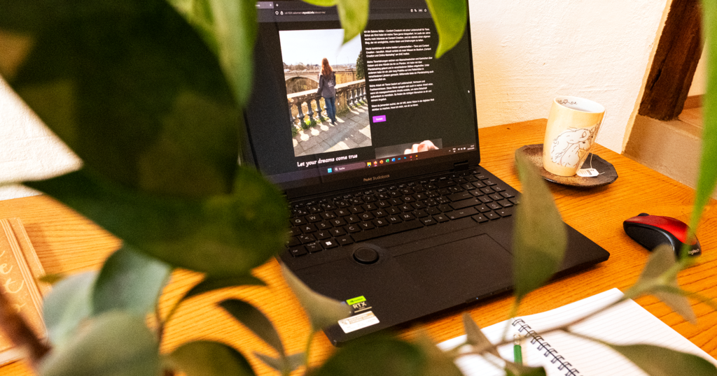 Laptop auf Holztisch hinter grüner Pflanze, daneben Tasse Tee und Notizbuch.