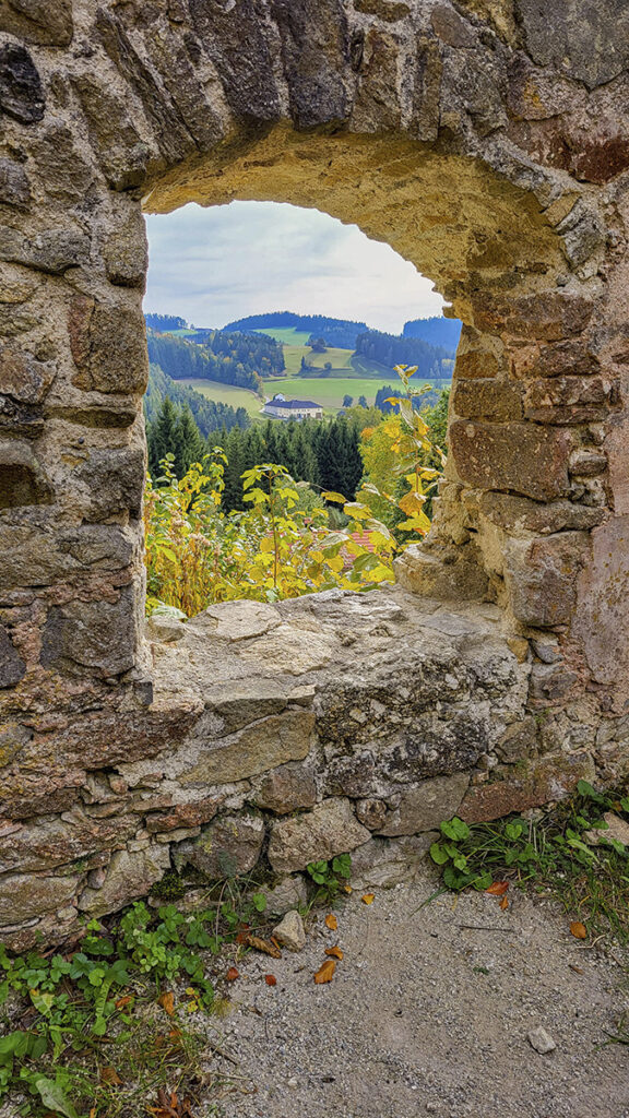 Blick durch ein altes Steinfenster auf Hügel, Wälder und ein abgelegenes Haus.
