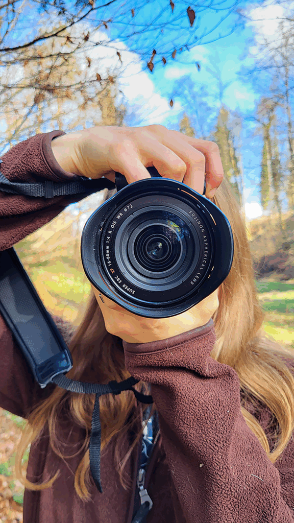 Person hält Fujifilm-Kamera im Wald, blauer Himmel und Herbstblätter im Hintergrund.